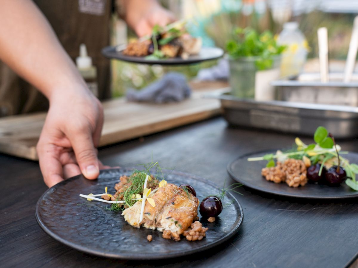 A person is serving plated gourmet dishes with garnishes, cherries, and unique presentation on a dark table surface.