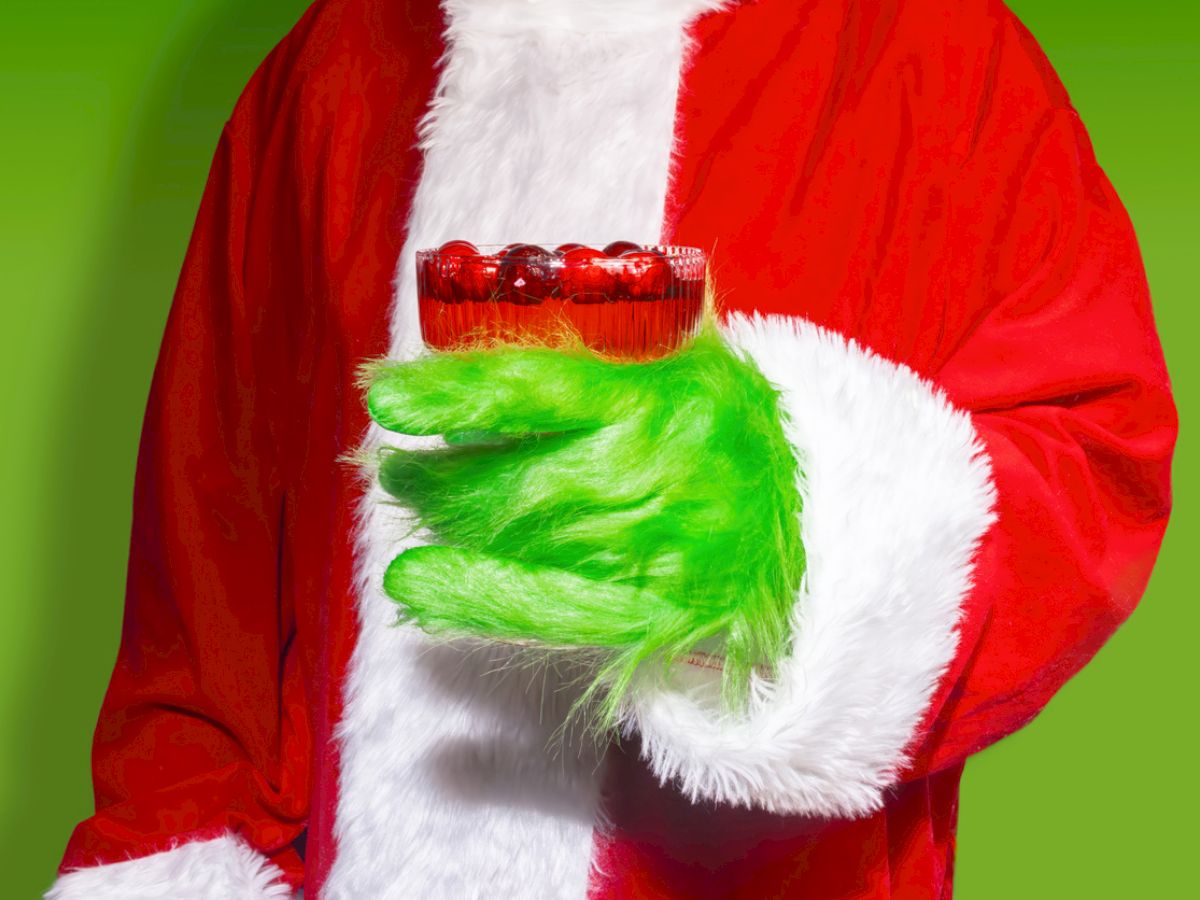 A person in a Santa suit with green, furry gloves holding a red glass against a green background.