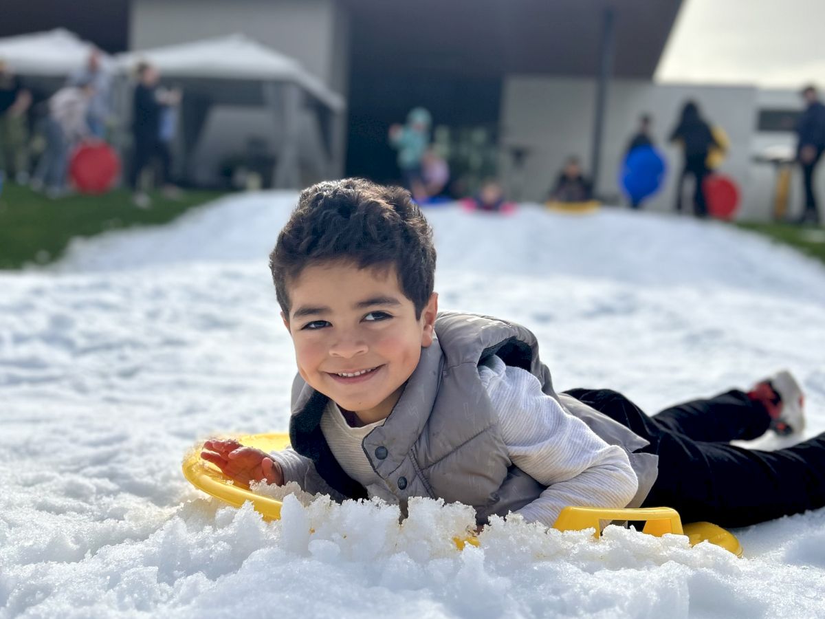 A child is lying on snow, smiling, with a house and several people in the background.