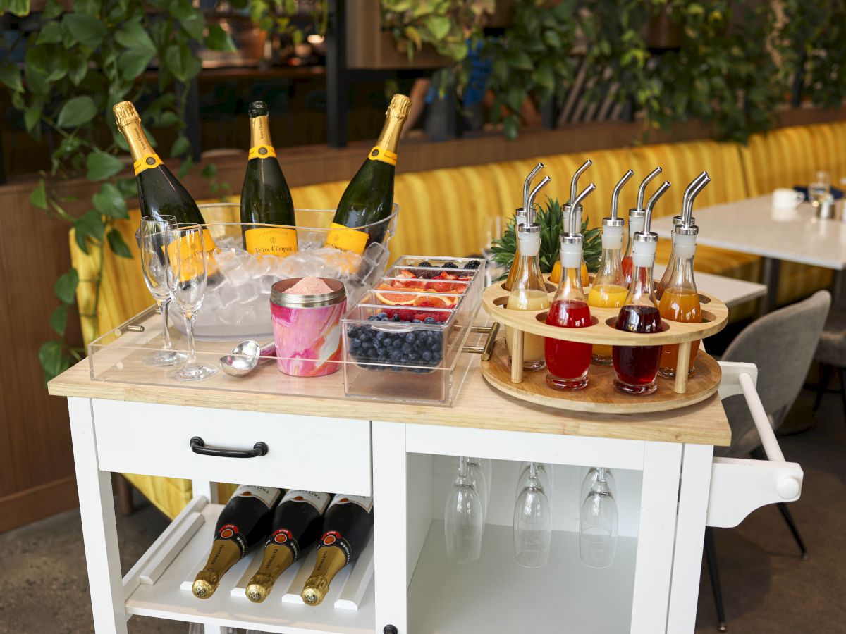 A drinks cart with champagne, glasses, fruits, and syrups is set up in a dining area with yellow seating.