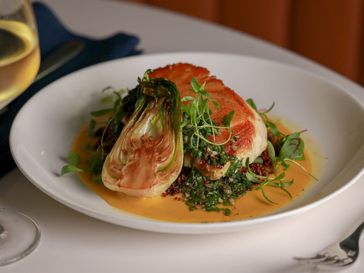 A gourmet dish with a seared fish fillet atop greens and a yellow sauce, served with a glass of white wine on a white table.