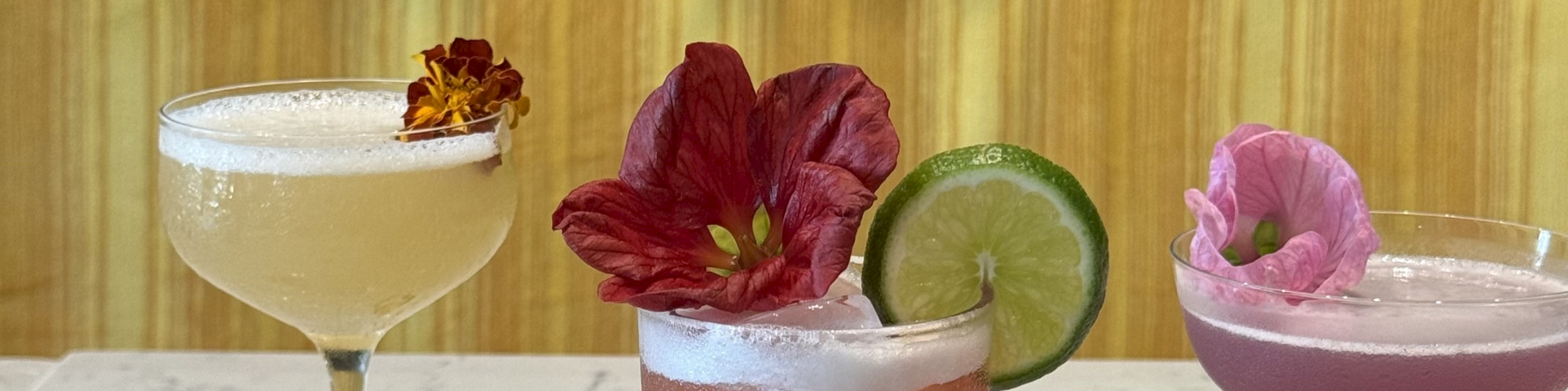 Three colorful cocktails on a wooden bar: pale yellow, pink with lime slice, and a purple drink, each garnished with tropical flowers.