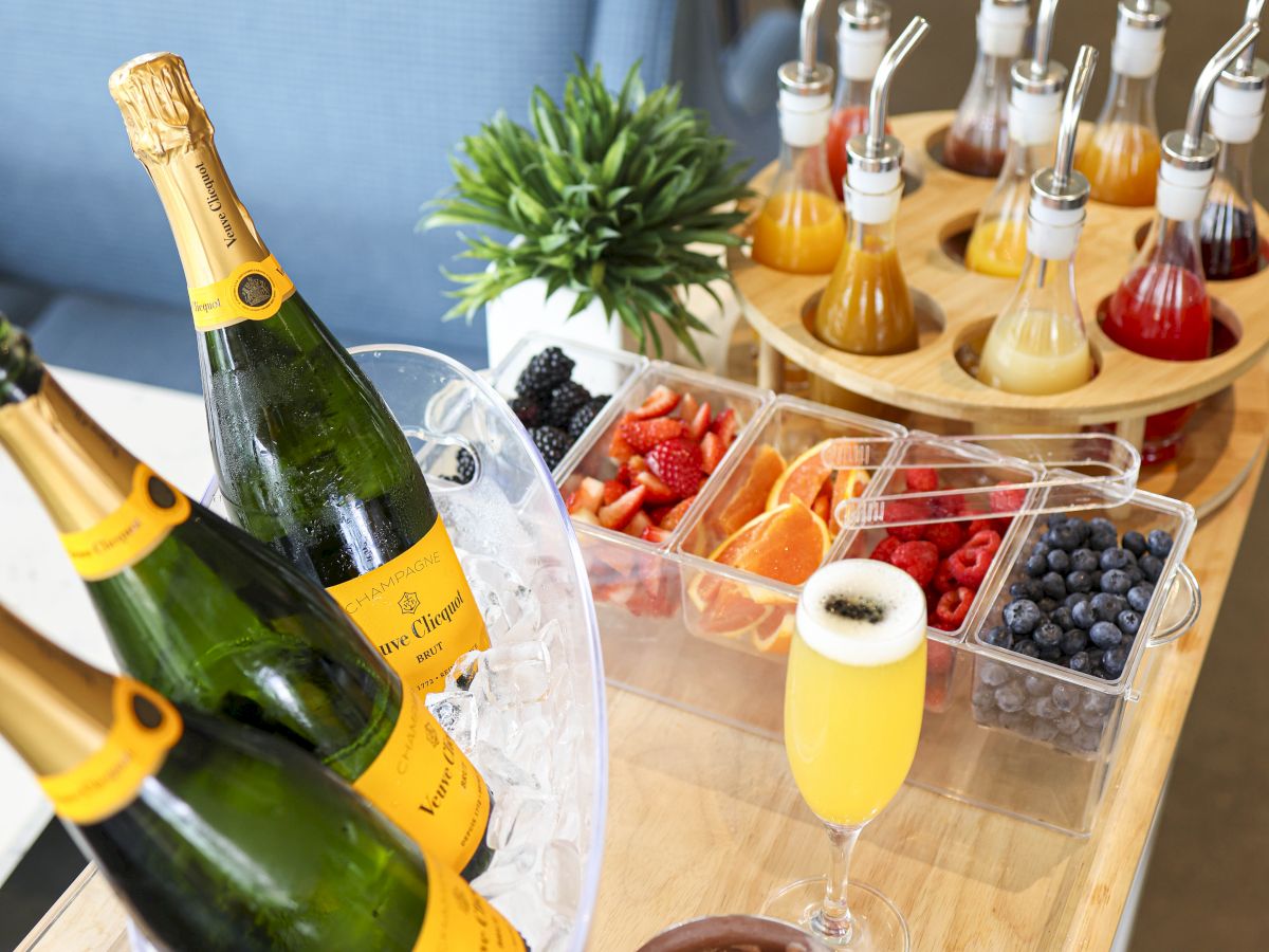 The image shows champagne bottles on ice, fruit garnishes, and syrups on a table, resembling a mimosa or cocktail setup.