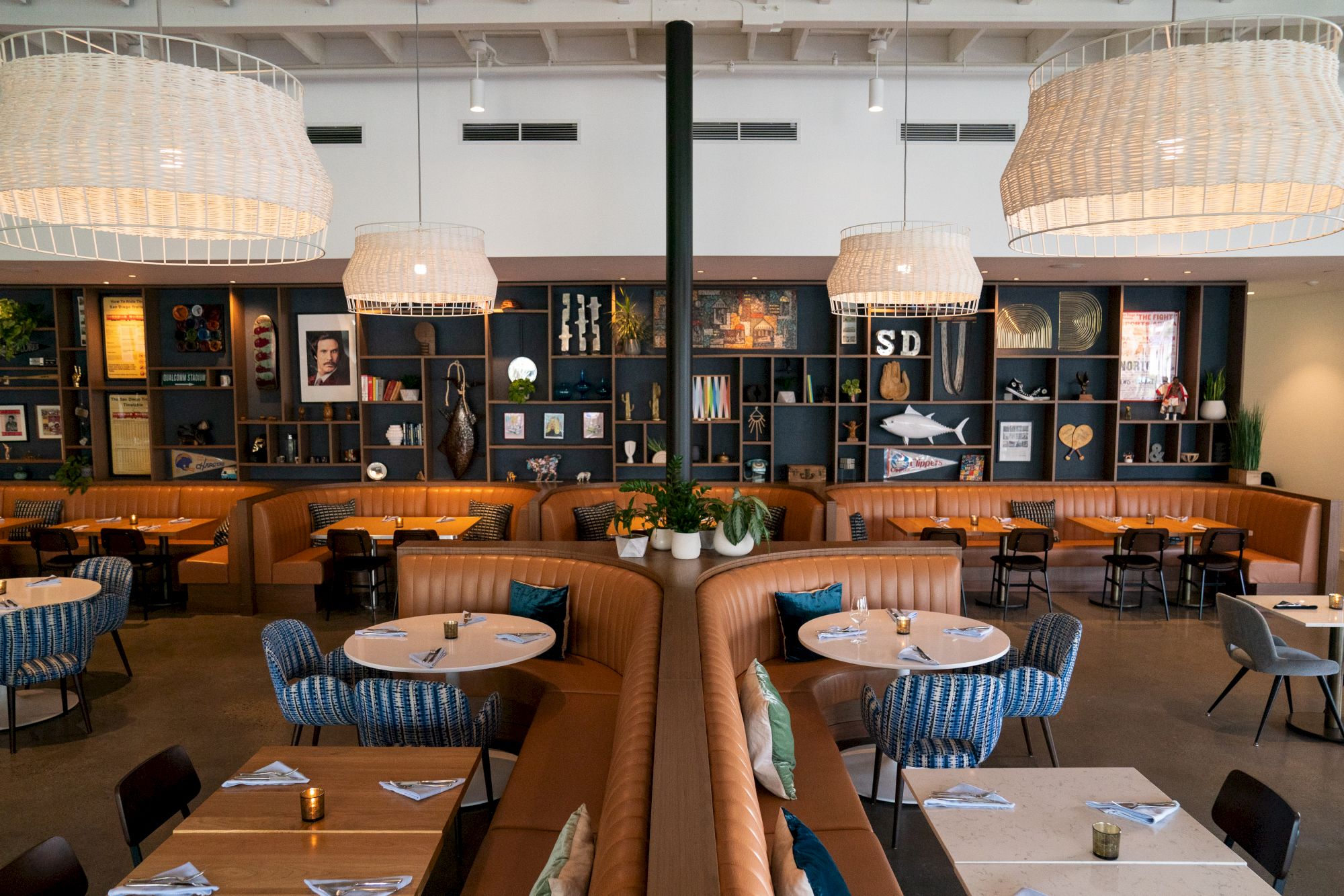 A stylish restaurant interior with pendant lighting, round tables, and cushioned booths. Decorative shelves display various items and art.