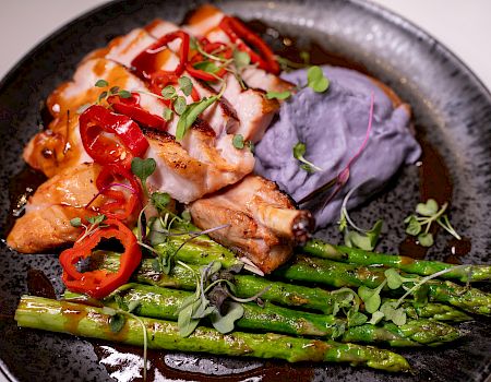 The image shows a plate with grilled salmon, asparagus, purple mashed potatoes, topped with sliced red peppers and microgreens.