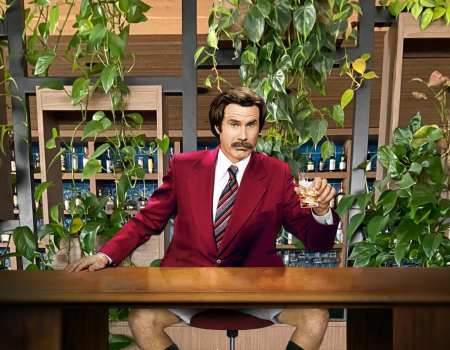 A man in a red jacket, holding a glass, sits at a desk surrounded by plants, with a humorous twist of wearing no pants.
