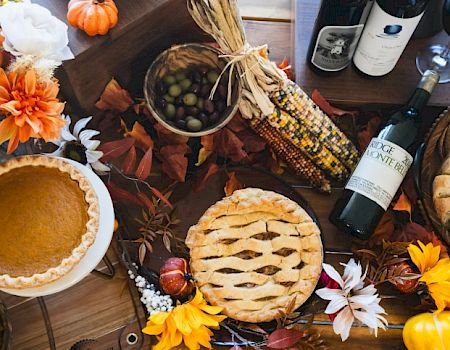 A festive table with pies, bread, corn, grapes, wine, and autumn decorations including pumpkins and flowers.