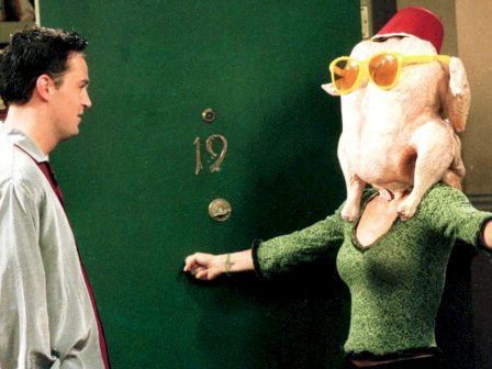 A person with a turkey on their head wearing glasses and a hat, while another person looks on, standing in front of a door numbered 19.