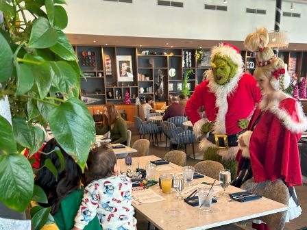 A child and adult at a restaurant table are visited by people in Grinch-themed costumes, decorated for the holiday season.