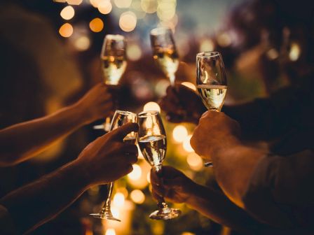 People are raising champagne glasses in a celebratory toast, surrounded by warm, glowing lights in a festive atmosphere.
