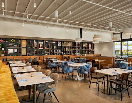 A bright modern cafe with wooden tables, blue and beige chairs, a long yellow bench, a chalkboard wall, and shelves behind a bar area.
