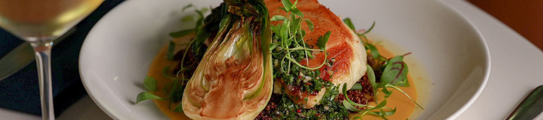 A plated gourmet dish with seared salmon on greens, nestled in a creamy sauce, served with a glass of white wine.