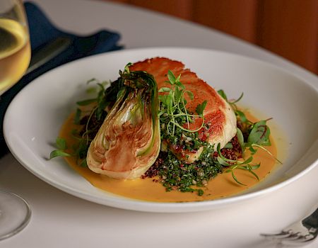 A plated gourmet dish with seared salmon on greens, nestled in a creamy sauce, served with a glass of white wine.