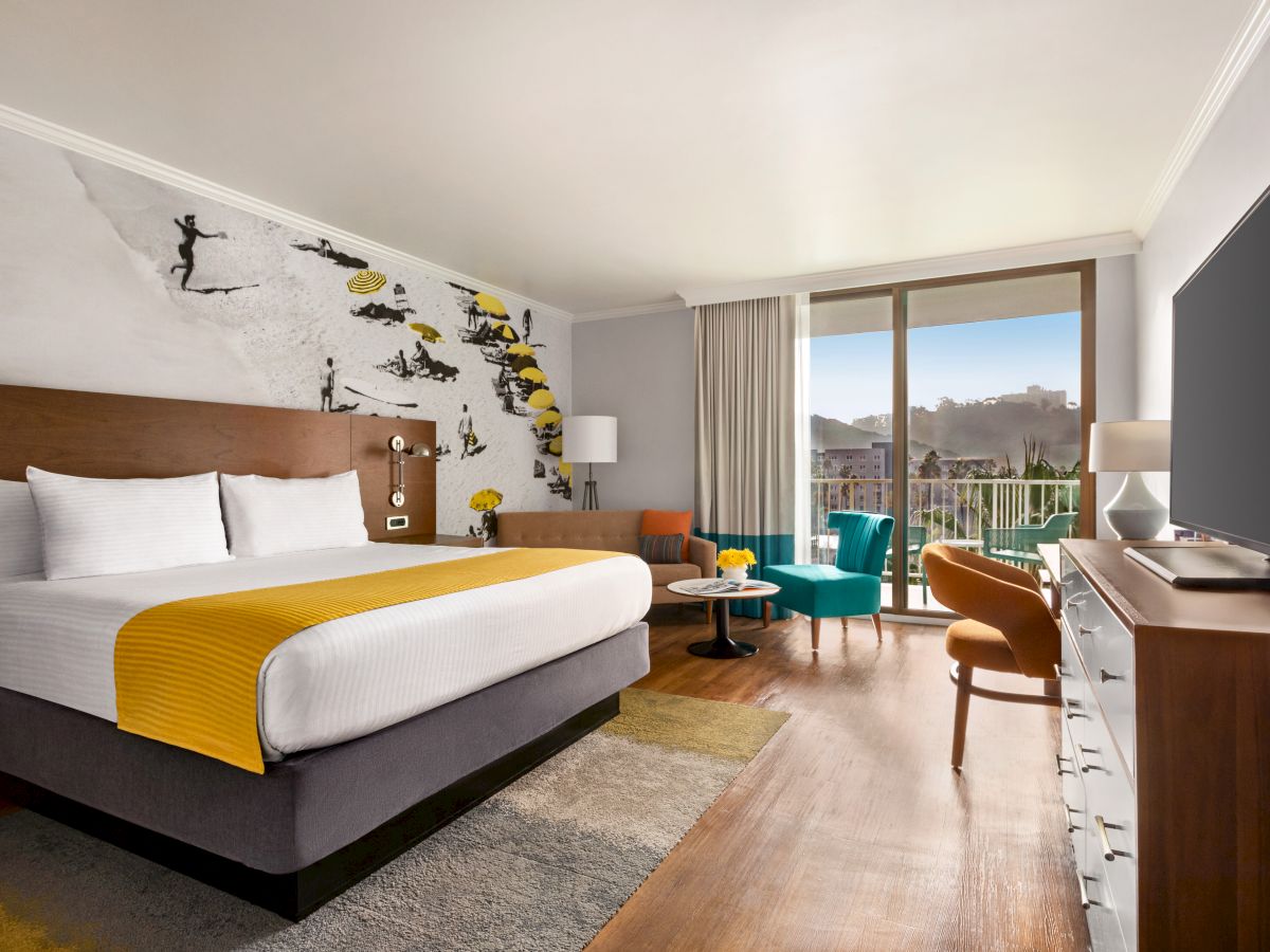 A stylish hotel room with a king bed, mustard throw, wall art, a sitting area by a balcony, and a flat-screen TV across from a wooden dresser.