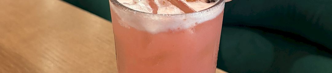 A pink lemonade cocktail with a strawberry and lemon garnish, a black straw, on a wooden table in a stylish lounge.