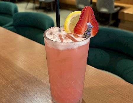 A pink lemonade cocktail with a strawberry and lemon garnish, a black straw, on a wooden table in a stylish lounge.