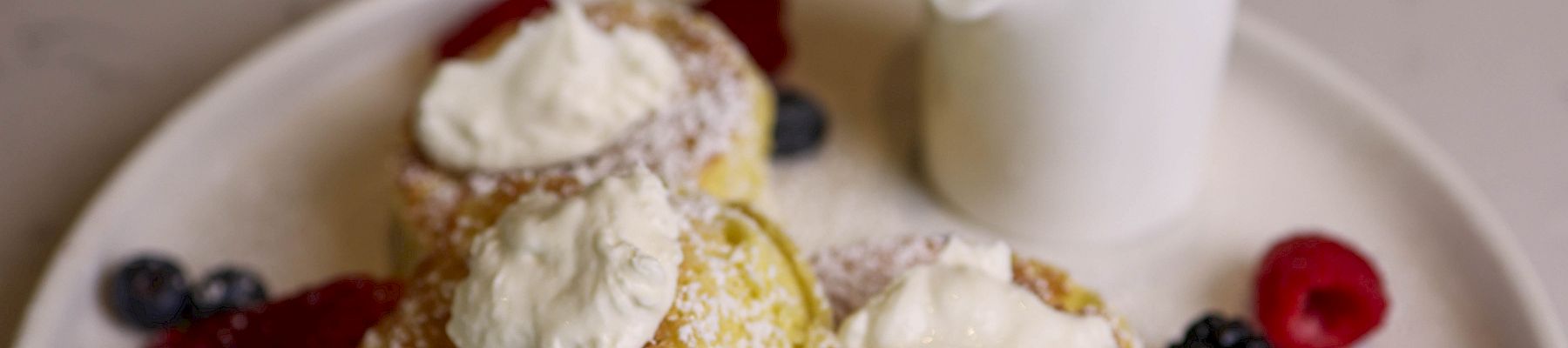 Two fluffy lemon pancakes topped with whipped cream, dusted with sugar, served with a side jug of syrup and fresh berries on a white plate.
