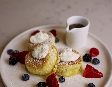 Two fluffy lemon pancakes topped with whipped cream, dusted with sugar, served with a side jug of syrup and fresh berries on a white plate.