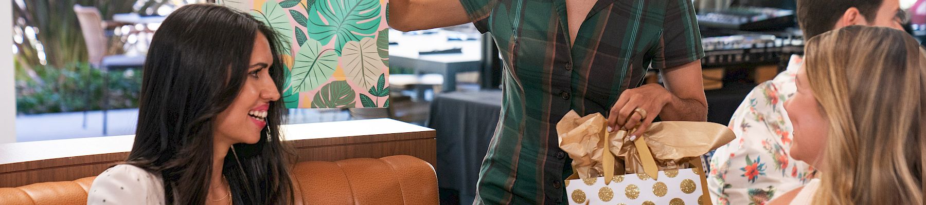 Two friends are dining; one brings a gift bag and a box of pastries, while the other accepts a paper bag, smiling over mimosas.