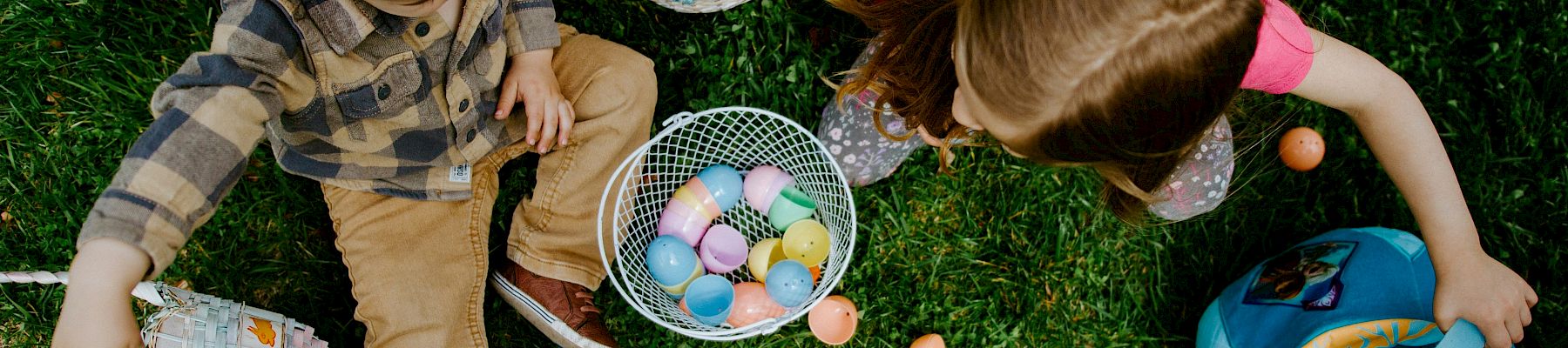 Two children with baskets sit on grass collecting colorful Easter eggs; a blue bucket sits nearby, and pastel eggs are scattered.