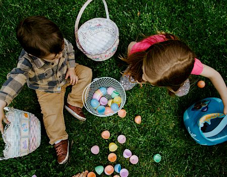 Two children with baskets sit on grass collecting colorful Easter eggs; a blue bucket sits nearby, and pastel eggs are scattered.