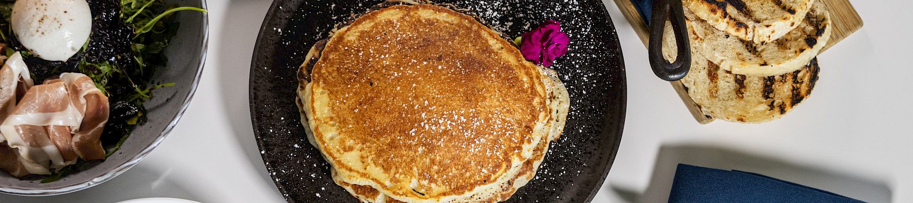 A top-down brunch spread: pancakes on a black plate, avocado toast bowls, crispy bacon, greens, pastries, grilled bread, small bowls, and a glass of orange juice all together.
