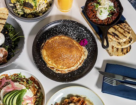 A top-down brunch spread: pancakes on a black plate, avocado toast bowls, crispy bacon, greens, pastries, grilled bread, small bowls, and a glass of orange juice all together.