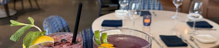 Three colorful cocktails on a wooden tray in a chic restaurant, with empty tables and bookshelves in the background ending with a period.