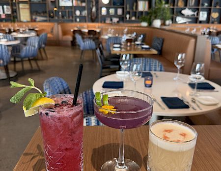 Three colorful cocktails on a wooden tray in a chic restaurant, with empty tables and bookshelves in the background ending with a period.