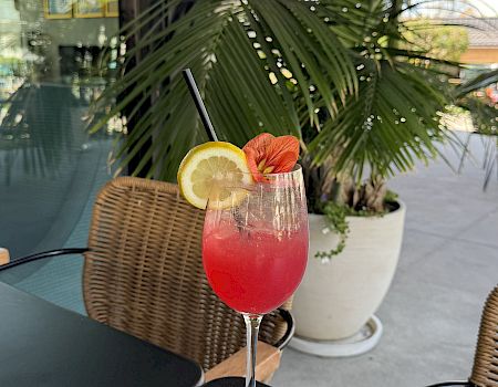 A red tropical cocktail with a lemon wheel, strawberry slice, and straw on a patio table by wicker chairs and large green plants.