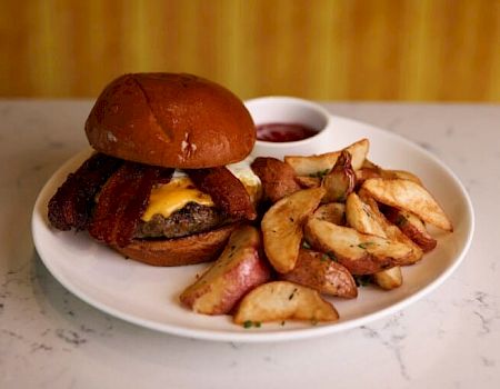 A cheeseburger with bacon and a side of potato wedges on a white plate, plus a small cup of dipping sauce, set on a light counter.