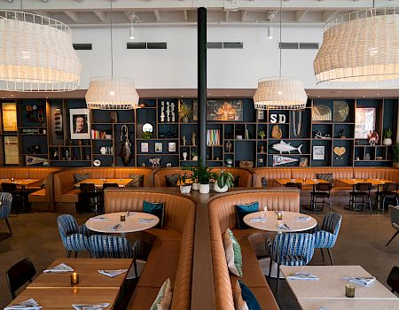 A stylish restaurant with booth seating, round tables, blue chairs, warm lighting, and a long wall of shelves and framed art in the background.