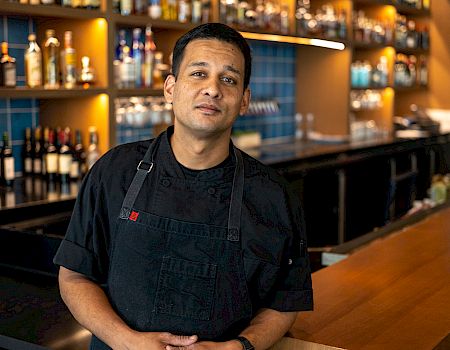 A chef stands behind a bar or counter in a kitchen or restaurant, wearing a black apron, with shelves of bottles in the background.