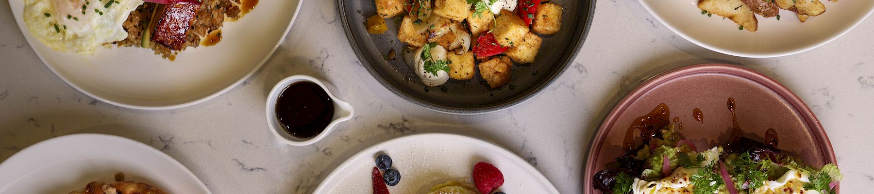 A colorful brunch spread: sunny-side eggs, pancakes with berries, a burrito bowl, skillet veggies, burgers with fries, waffles, and assorted syrups and sauces, delicious.