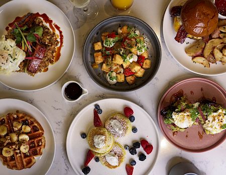 A colorful brunch spread: sunny-side eggs, pancakes with berries, a burrito bowl, skillet veggies, burgers with fries, waffles, and assorted syrups and sauces, delicious.