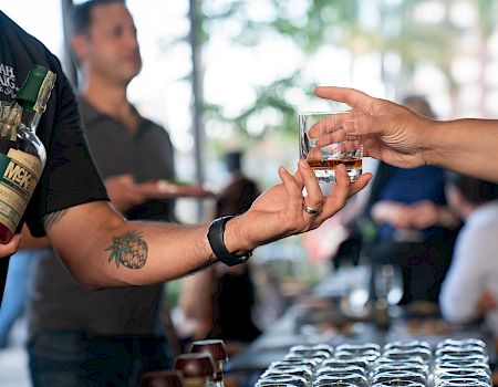 Two people exchanging a drink at a bar: a bartender pours whiskey into a glass while another hand reaches to take it, with bottles and glasses on the counter.