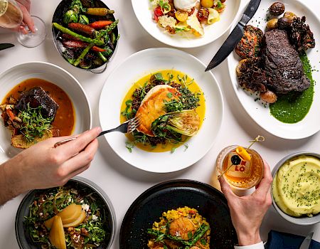 A top-down spread of gourmet plates with steak, roasted vegetables, pasta, and sauces, ready for a shared meal.