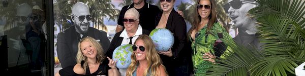 A group of friends in casual beachwear pose for a photo in front of a &ldquo;Dali&rsquo;s Arlo&rdquo; banner with palm trees.