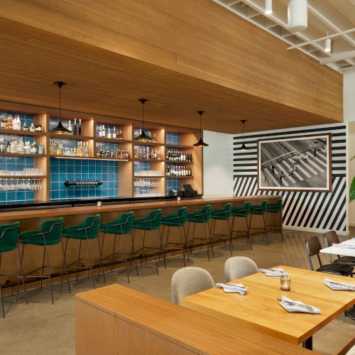 Modern bar interior with a long wooden counter, hanging lights, shelves of bottles, and green stools, plus a framed wall photo.