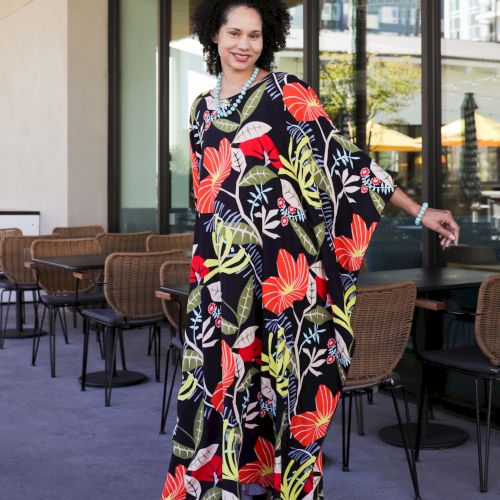 A woman in a bold floral dress with a pearl necklace smiles outside at a modern caf&eacute; with outdoor chairs and glass windows.