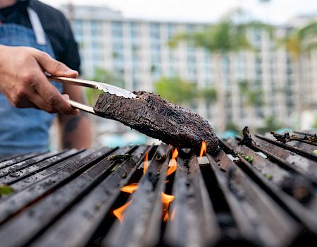 A person grills a charred piece of meat over a flaming outdoor charcoal fire using tongs in front of a building backdrop.
