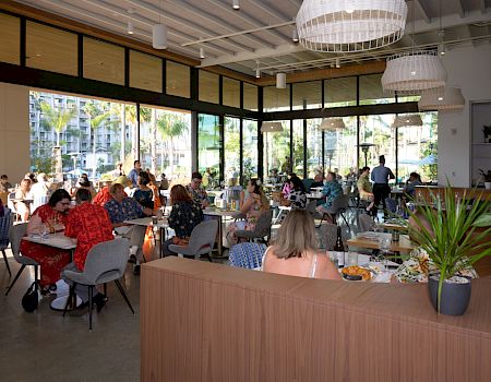 A bright dining hall packed with families, tables set with food and drinks, overlooking palm-filled resort views.