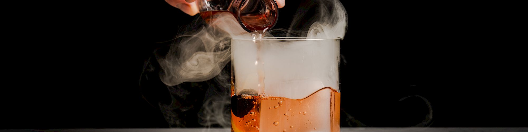A hand pours amber liquor from a bottle into a glass, with billowing smoke rising against a dark background.