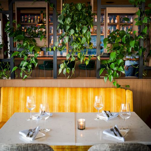 A cozy restaurant table set with wine glasses, lit candles, and hanging green plants framed on a wooden wall, for dining.