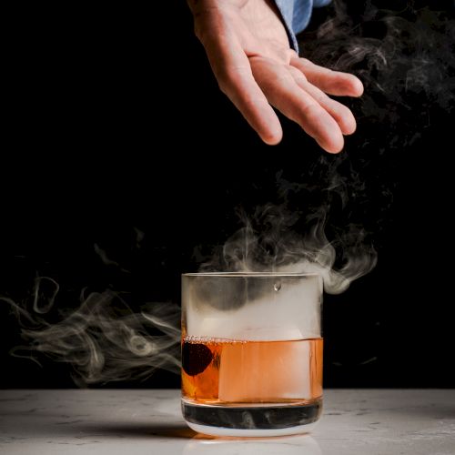 A hand hovers over an on-the-rocks whiskey glass as smoke billows in a dark, dramatic setting.