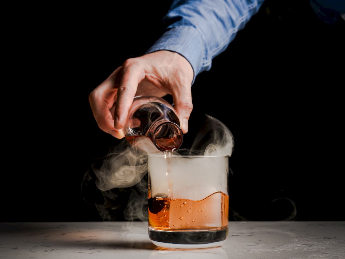 A hand pours amber liquor from a bottle into an ice-filled tumbler as smoke billows in a dark scene.