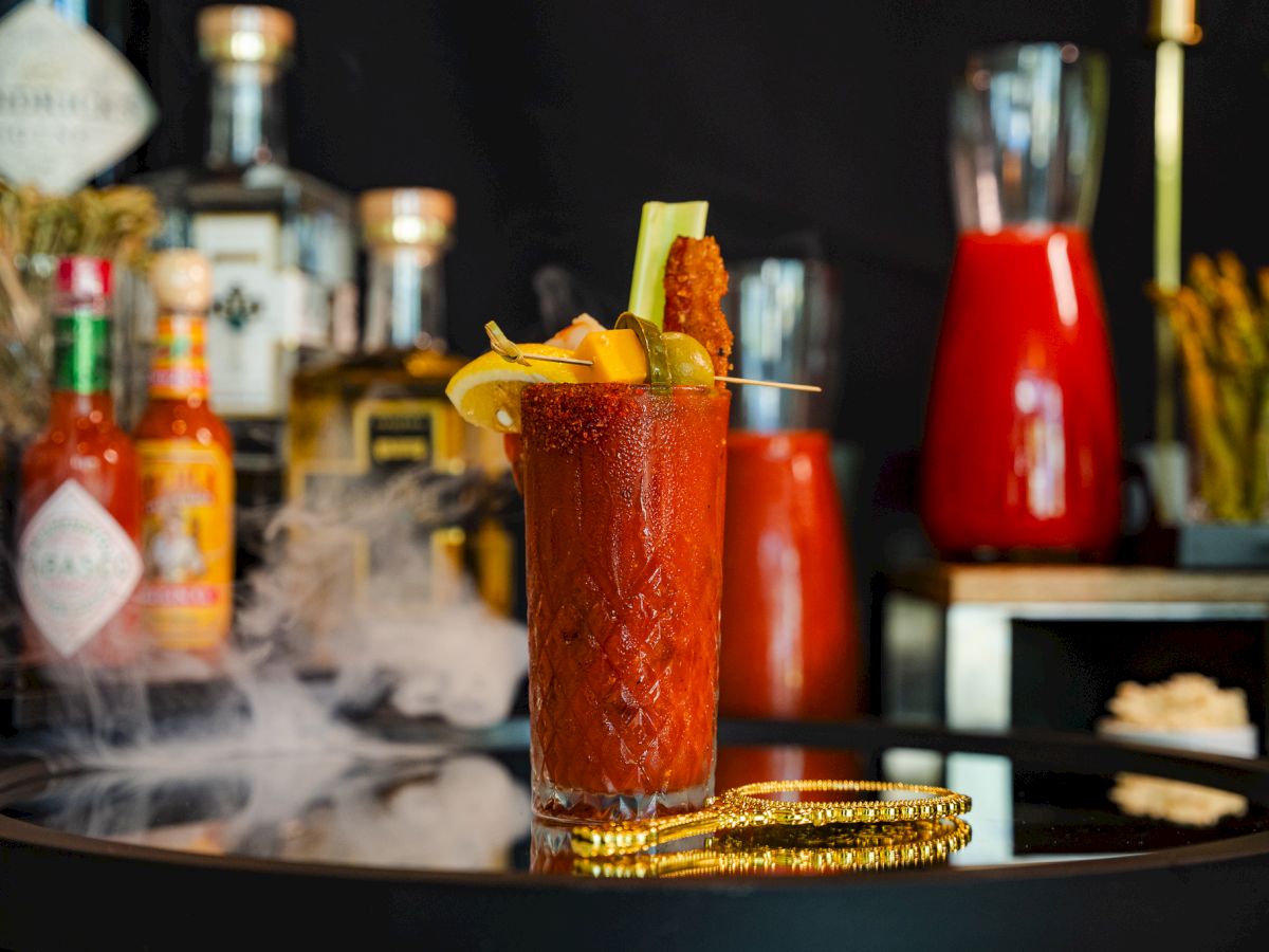 A tall red cocktail in a glass, garnished with an olive and skewer, sits on a bar with bottles blurred behind it.