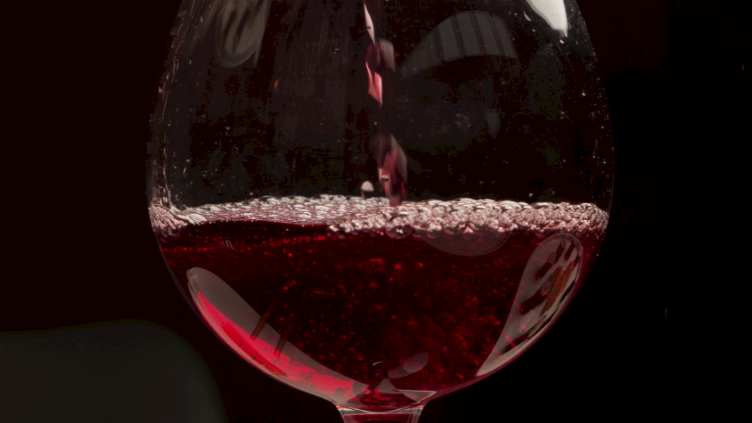 A stream of red wine is pouring into a glass, forming bubbles and splashes against a dark background.