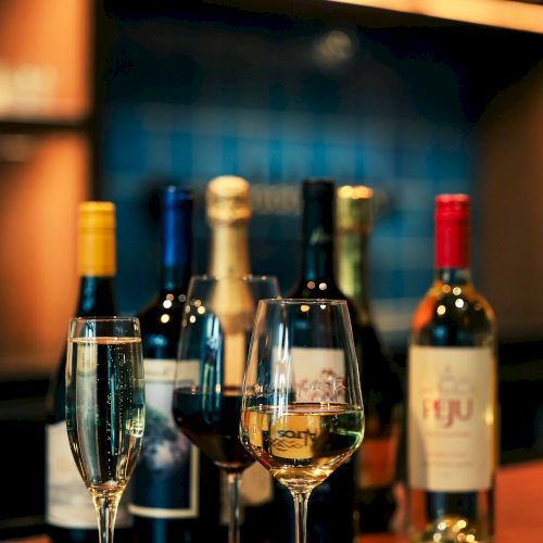 A cozy bar scene with bottles and two filled wine glasses under warm lights, blurred background.