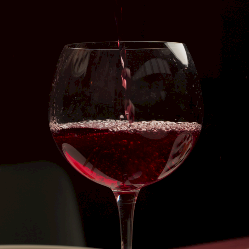 A stream of red wine pours into a glass, filling it with deep ruby color and bubbles.